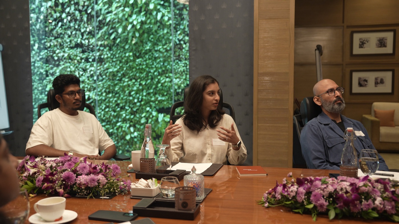 Three participants seated at the table, one speaking, a green living wall behind them.