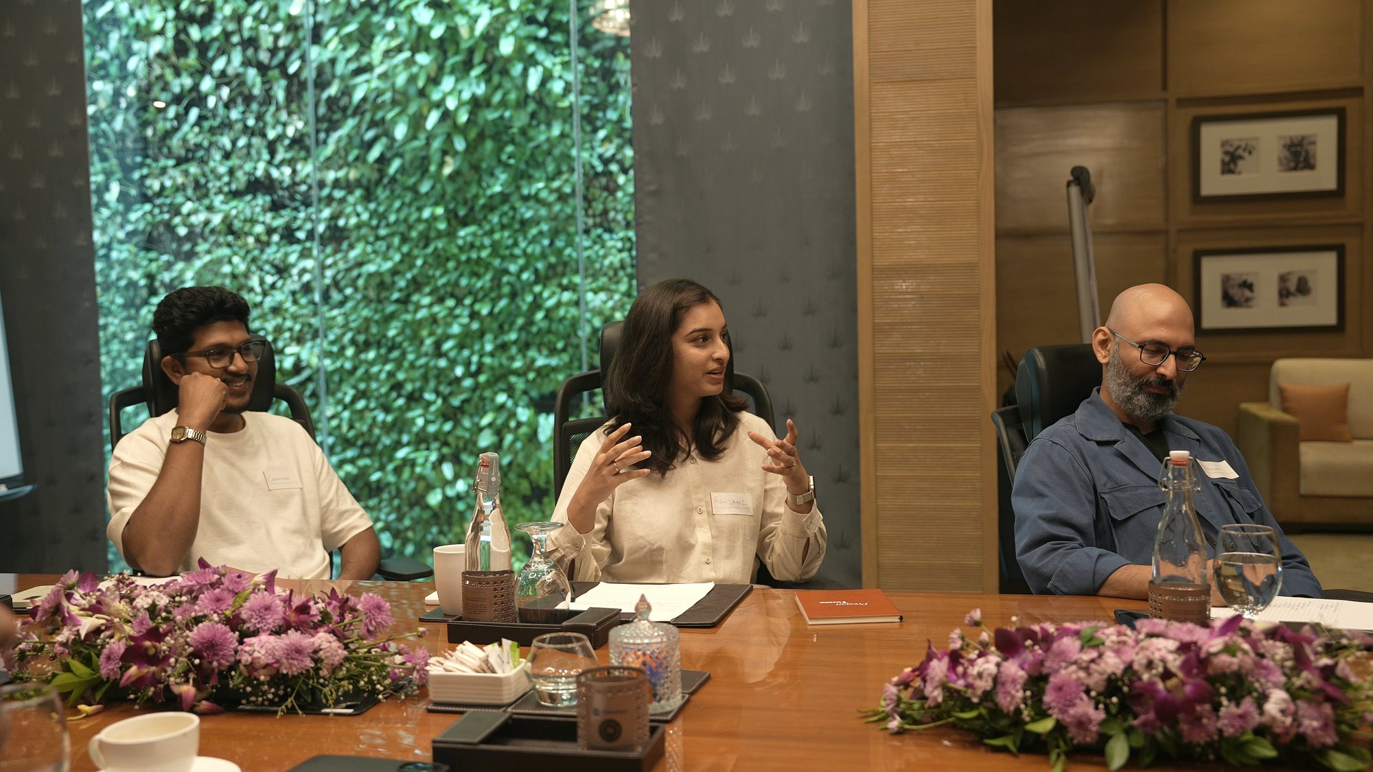 Three participants at the boardroom table, one mid-story, a green living wall behind them.